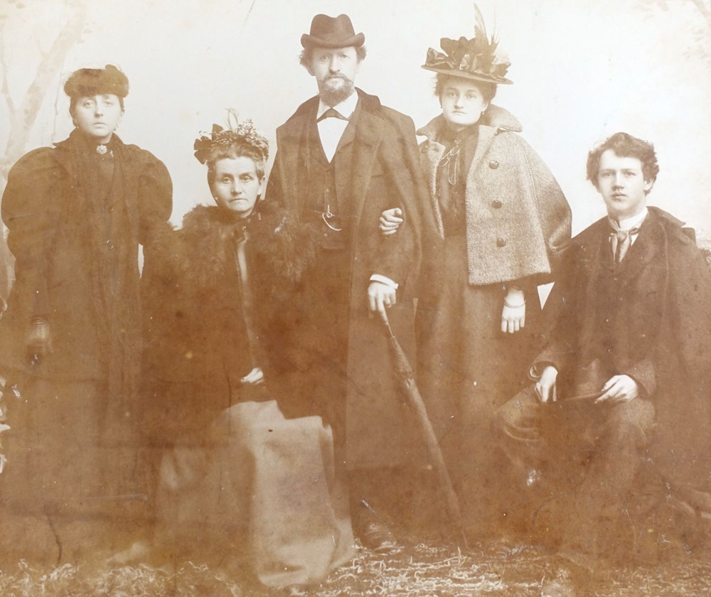 Eine historische Schwarz-Weiß-Fotografie zeigt eine Gruppe von fünf Personen in formeller Kleidung, darunter ein Mann mit Hut und eine Frau mit einem auffälligen Hut.