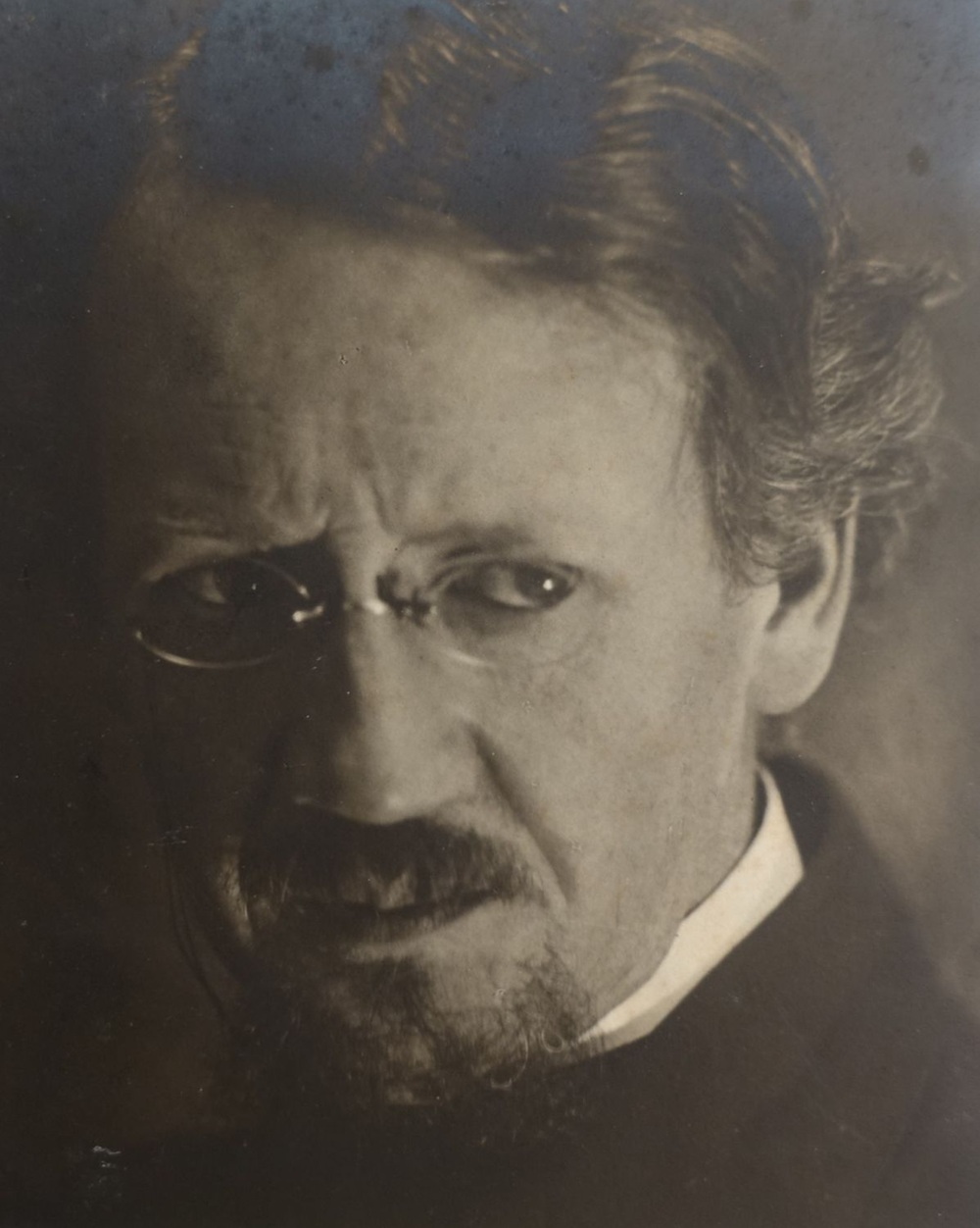 Ein Mann mit Brille bart und oberlippenbart und ernstem Gesichtsausdruck, um 1920. Es ist der Schweizer Künstler Fritz Gilsi.