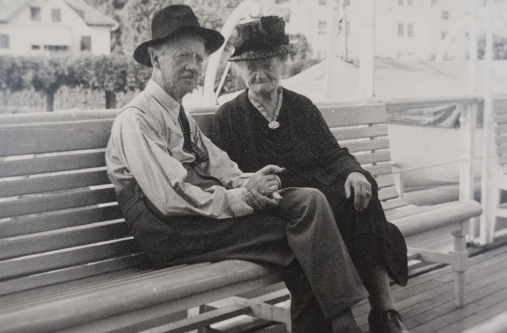 Ein älteres Paar sitzt eng beieinander auf einer Park-Bank, beide in Sonntagsgewand mit Hut. Der Mann ist der Schweizer Maler Fritz Gilsi.